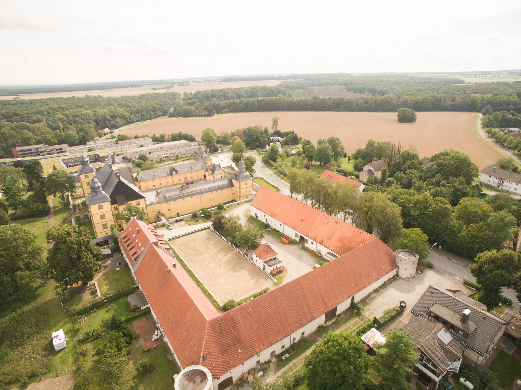Luftaufnahme von Eringerfeld: das gelbe Schloss mit seinen Ecktürmen, davor die langen Wirtschaftsgebäude mit roten Ziegeldächern um den Reitplatz, dahinter Wald und abgeerntete Felder