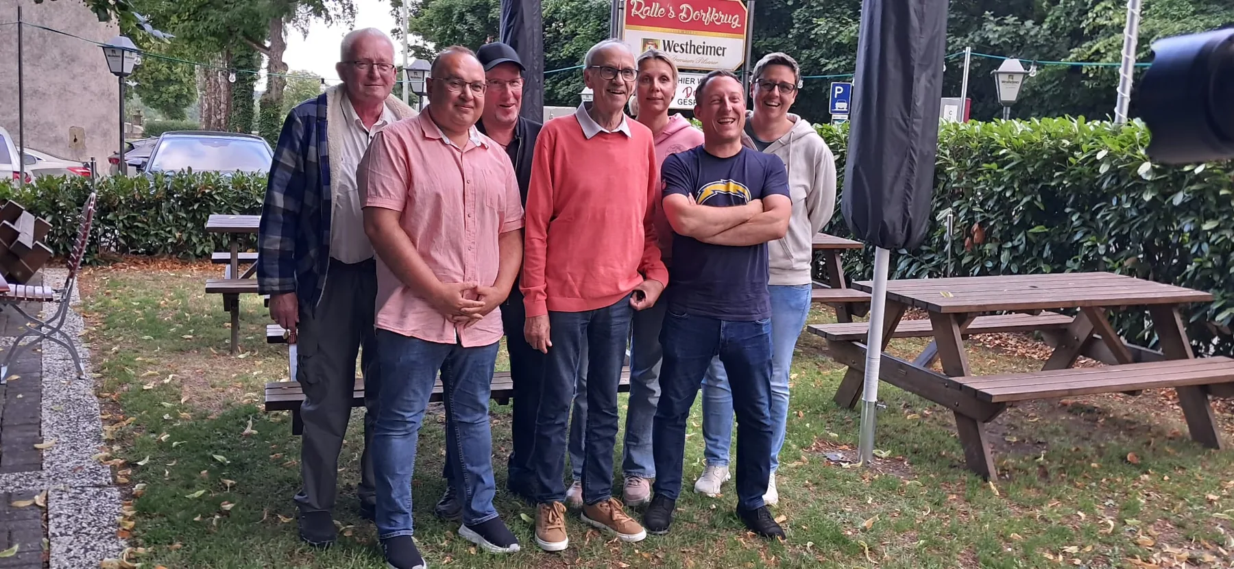 Mitglieder des Heimatvereins nebeneinander im Garten eines Dorfgasthauses, dahinter Biertischgarnituren und eine Hecke